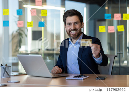 Confident businessman in formal attire holding credit card at office desk, working on laptop, showing happiness and satisfaction. Colorful sticky notes on glass wall suggesting planning or 117902662