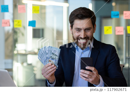 Businessman expresses happiness holding cash and checking smartphone. Man dressed in formal suit shows achievement and financial success, depicting concepts of wealth, communication, and business. 117902671