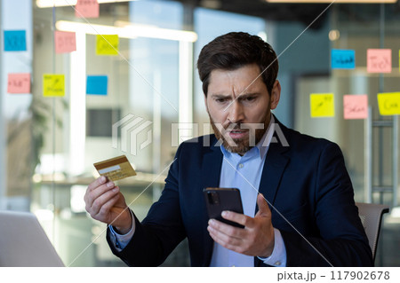 Concerned businessman in suit holding credit card and phone, looking at phone screen with worried expression, dealing with online banking or payment problem in modern office setting Concerned businessman in suit holding credit card and phone, looking at phone screen with worried expression, dealing with online banking or payment problem in modern office setting 117902678