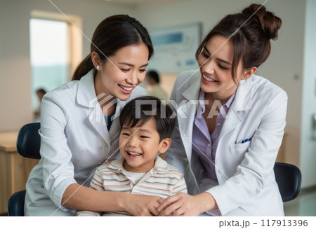 Two female dentists smile with a young child patient in a modern dental clinic, creating a supportive and friendly atmosphere. The scene emphasizes trust and comfort in pediatric dental care. 117913396