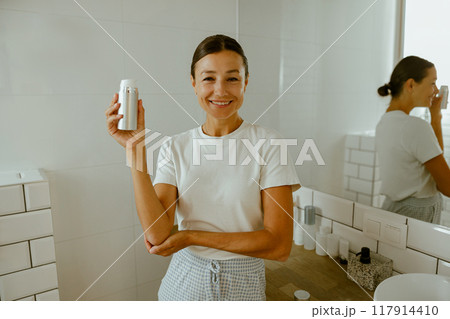 A Joyful Smiling Woman Elegantly Showcasing a Skincare Product in a Modern Bathroom Setting A Joyful Smiling Woman Elegantly Showcasing a Skincare Product in a Modern Bathroom Setting 117914410