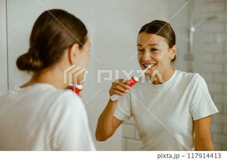 A cheerful and confident woman smiling while brushing her teeth in a contemporary bathroom 117914413