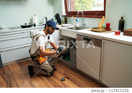 Professional male plumber in uniform sitting near kitchen sink and writing on clipboard in kitchen 117915258