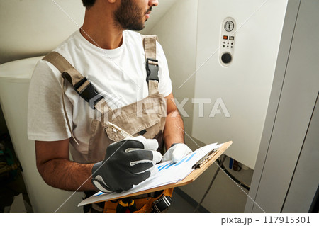 Professional male handyman in uniform is checking gas boiler at home and making notes in tablet  117915301