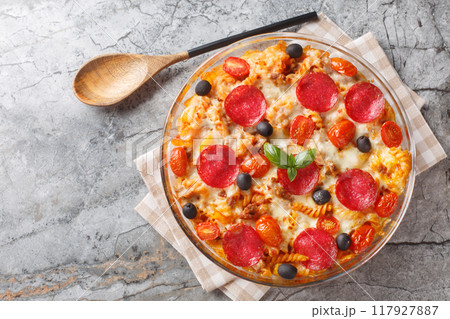 Pasta pizza casserole with cheese, fried ground beef, salami sausage, peppers and olives with marinara sauce close-up in glass baking dish. Horizontal top view 117927887
