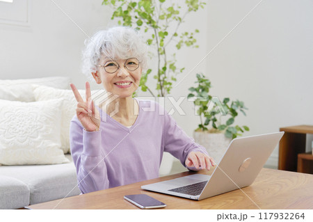 ノートパソコンの前でピースサインするシニア女性　リビングルーム 117932264
