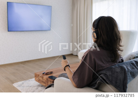 Boy with a long hair watch TV and use wireless headphones. Home alone. Boy with a long hair watch TV and use wireless headphones. Home alone. 117932785