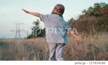 Happy boy in aviator pilot helmet runs as airplane on nature meadow, outside city. Kids dream is to 117934586