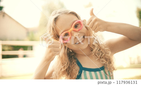 Smiling little blonde girl wearing swimming goggles shows thumbs up sign at outdoor swimming pool. Happy summer vacation time 117936131