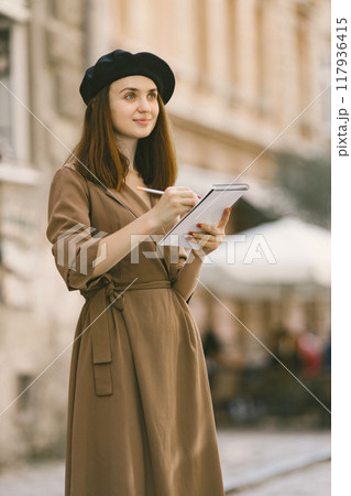 Female painter painting while standing in a city center. A woman wearing khaki dress and a black hat. Girl drawing a sketch in summer on a street. Female painter painting while standing in a city center. A woman wearing khaki dress and a black hat. Girl drawing a sketch in summer on a street. 117936415