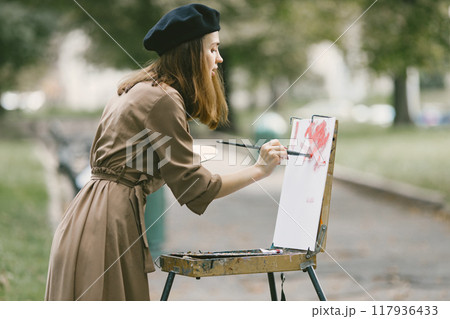 Female painter painting in the park. A woman wearing khaki dress and a black hat. Girl drawing a picture on an easel in summer on nature. 117936433