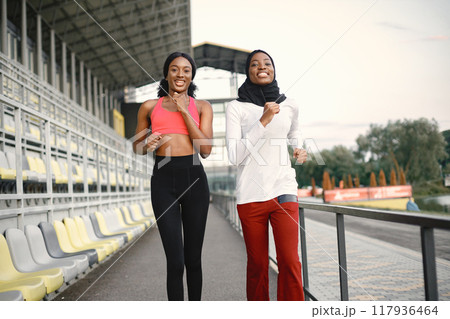 Two women running on a stadium. Muslim woman in hijab wearing white shirt and red trousers. Brunette woman wearing rose top and black leggins. 117936464