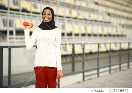 Girl working out outdoors on a stadium. Muslim woman wearing white shirt and red trousers. Girl doing excercises with a dumbbells. 117936473