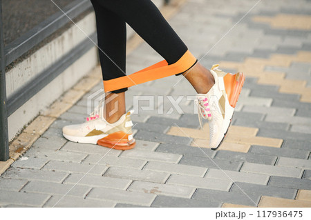 Close up of legs and equipment for practicing yoga in the studio, band for doing stretching. Afro american woman doing workout on a stadium. Woman wearing white and orange sneakers. Close up of legs and equipment for practicing yoga in the studio, band for doing stretching. Afro american woman doing workout on a stadium. Woman wearing white and orange sneakers. 117936475