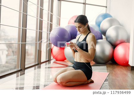 Fitness girl working out at gym with big window. Girl wearing black leggins and sport top. Young woman sitting on a yoga mat and using a wireless earphones. Fitness girl working out at gym with big window. Girl wearing black leggins and sport top. Young woman sitting on a yoga mat and using a wireless earphones. 117936499