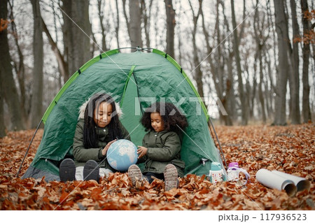 Beautiful little black girls in tent camping in the forest. Two little sisters sitting in a tent in autumn forest and looking on a globe. Black girls wearing khaki coats. 117936523
