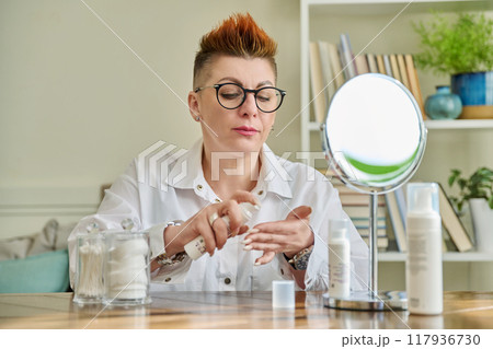 Middle aged woman looking in mirror, using face creams, cosmetic care products 117936730