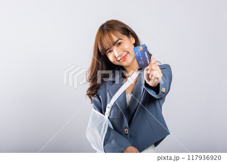 Smiling woman after accident with a broken arm, confidently using a splint. Paying medical bills by credit card. Happy Asian female with sling support isolated on white background. Injury is evident. Smiling woman after accident with a broken arm, confidently using a splint. Paying medical bills by credit card. Happy Asian female with sling support isolated on white background. Injury is evident. 117936920