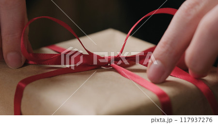 closeup shot of young female hands unties red ribbon bow on craft paper gift box closeup shot of young female hands unties red ribbon bow on craft paper gift box 117937276
