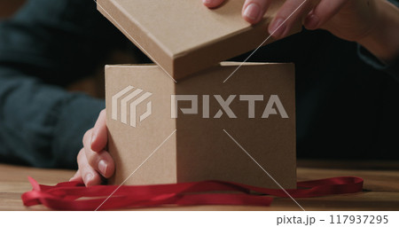 closeup shot of young female hands opens craft paper gift box with red ribbon bow closeup shot of young female hands opens craft paper gift box with red ribbon bow 117937295