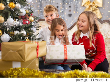 Happy brother with two sisters are sitting on sofa together with gift boxes next to the new year tree 117939921