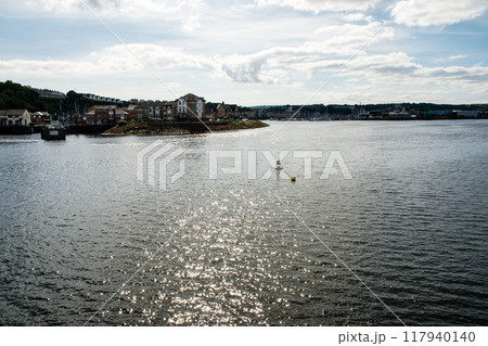 日差しが水面に反射してまぶしい入江と遠くに見えるヨットハーバーとレンガ造りの建物　南ウェールズにて 117940140