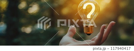 Hand holding a glowing lightbulb with a question mark inside, blurred background of trees. Conceptual image representing inspiration, ideas, and curiosity. Hand holding a glowing lightbulb with a question mark inside, blurred background of trees. Conceptual image representing inspiration, ideas, and curiosity. 117940417