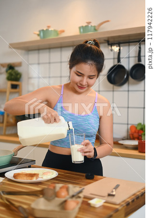 Attractive young woman pouring milk into a glass while standing in a kitchen Attractive young woman pouring milk into a glass while standing in a kitchen 117942226