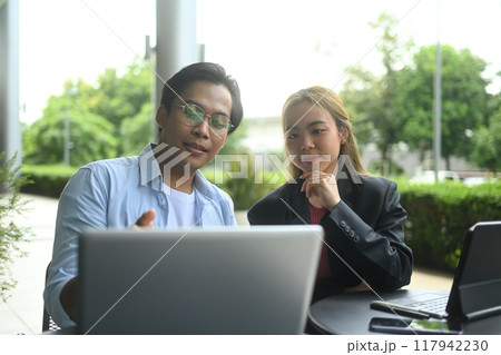 Two businesspeople discussing financial data on laptop while sitting in a modern office park 117942230