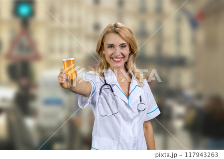 Happy blonde caucasian mature female doctor standing outdoors gives you can of pills. Blurred city street in the background. 117943263