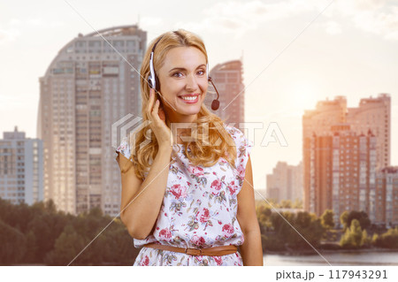 Portrait of happy mature blonde woman wearing headset outdoors. Customer support worker. Residential area buildings in the background. 117943291