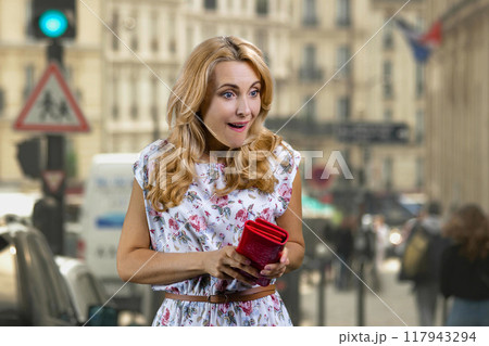 Portrait of happy mature surprised blonde woman taking money from her red wallet. City street in the background. 117943294