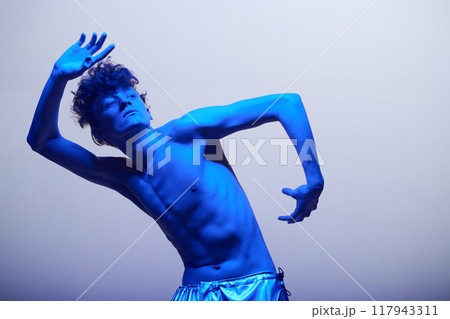 Shirtless man dancing and posing under bright blue lighting creating dramatic effect. Artistic, expressive performance partially obscured by shadows in minimalist setup Shirtless man dancing and posing under bright blue lighting creating dramatic effect. Artistic, expressive performance partially obscured by shadows in minimalist setup 117943311