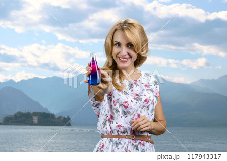 Portrait of happy blonde mature woman using perfume bottle. Sea and mountain landscape in the background. Portrait of happy blonde mature woman using perfume bottle. Sea and mountain landscape in the background. 117943317