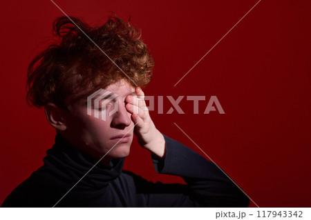 Young man lightly pressing his face with eyes closed while standing against vivid red background. His hair is tousled and he is wearing dark clothing Young man lightly pressing his face with eyes closed while standing against vivid red background. His hair is tousled and he is wearing dark clothing 117943342
