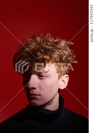 Thoughtful young man with curly hair against bright red background, casting shadow. Man turned slightly away from camera, creating a dramatic and introspective mood 117943343