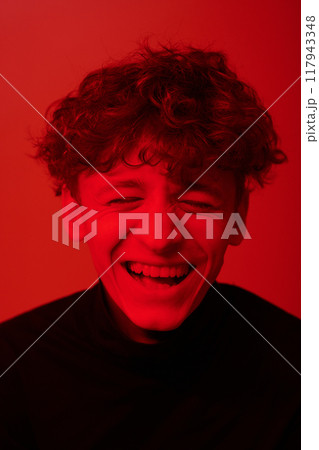 Young person captured mid-laughter, with vibrant red lighting creating an energetic ambiance. Curly hair and expressive face highlighting joyous emotion Young person captured mid-laughter, with vibrant red lighting creating an energetic ambiance. Curly hair and expressive face highlighting joyous emotion 117943348