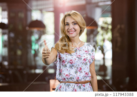 Portrait of charming mature blonde lady shows her thumb up. Restaurant indoors in the background. 117943388