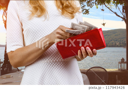 Close up female hands taking money from the red wallet. Sea or lake landscape in the background. Close up female hands taking money from the red wallet. Sea or lake landscape in the background. 117943426