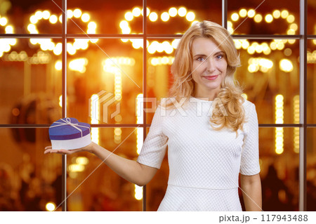 Happy mature blonde woman holding a blue heart shape gift box on her right palm. Christmas present for you. 117943488