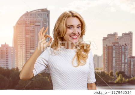 Portrait of a happy mature blond woman shows ok gesture sign. Residential area in the background. 117943534