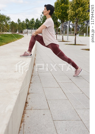 Side view of elderly woman in pink leggings stretching leg muscles on concrete bench while getting ready to run jogging outside in morning city park, copy space 117944265