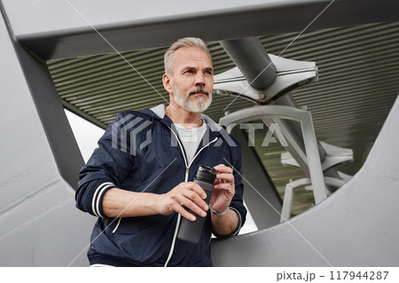 Medium shot of senior man with gray hair holding plastic bottle of water resting after daily jogging workout under shade pavilion at city park, copy space Medium shot of senior man with gray hair holding plastic bottle of water resting after daily jogging workout under shade pavilion at city park, copy space 117944287