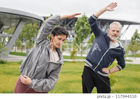 Medium shot of senior woman and man warming up side bending while working out together at morning city park Medium shot of senior woman and man warming up side bending while working out together at morning city park 117944310