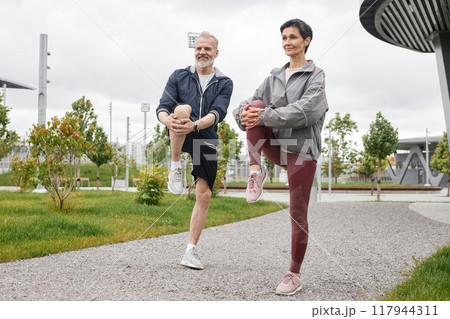 Full shot of smiling senior man and woman doing standing knee hugs, warming up and getting ready to run jogging at morning city park with gray sky Full shot of smiling senior man and woman doing standing knee hugs, warming up and getting ready to run jogging at morning city park with gray sky 117944311