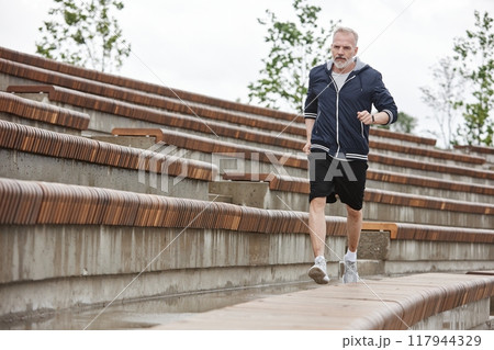 Full shot of fit elderly man in sportswear jogging along wooden benches of amphitheater working out regularly at city park, copy space Full shot of fit elderly man in sportswear jogging along wooden benches of amphitheater working out regularly at city park, copy space 117944329