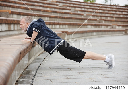 Side view of fit elderly man in sportswear doing push ups on on huge concrete steps of amphitheater working out regularly at city park, copy space 117944330