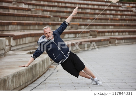 Full shot of fit elderly man in sportswear holding side plank while leaning on huge concrete steps of amphitheater working out core muscles regularly at city park, copy space Full shot of fit elderly man in sportswear holding side plank while leaning on huge concrete steps of amphitheater working out core muscles regularly at city park, copy space 117944331