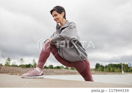 Smiling fit elderly woman in pink leggings stretching leg muscles in lunge position against cloudy sky in city park, copy space 117944339