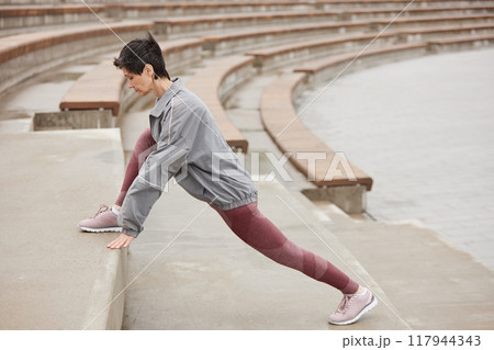 Side view of fit senior woman in pink sportswear stretching leg muscles on huge concrete steps of amphitheater getting ready to run jogging outside in city park, copy space Side view of fit senior woman in pink sportswear stretching leg muscles on huge concrete steps of amphitheater getting ready to run jogging outside in city park, copy space 117944343